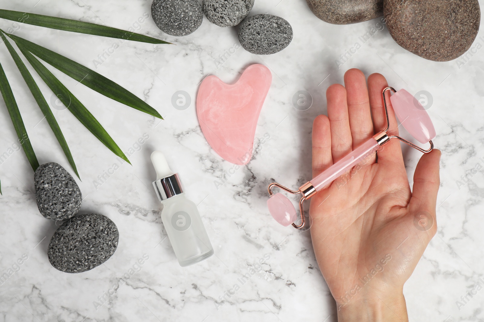 Photo of Woman with face roller, gua sha tool, cosmetic product and stones on white marble background, top view