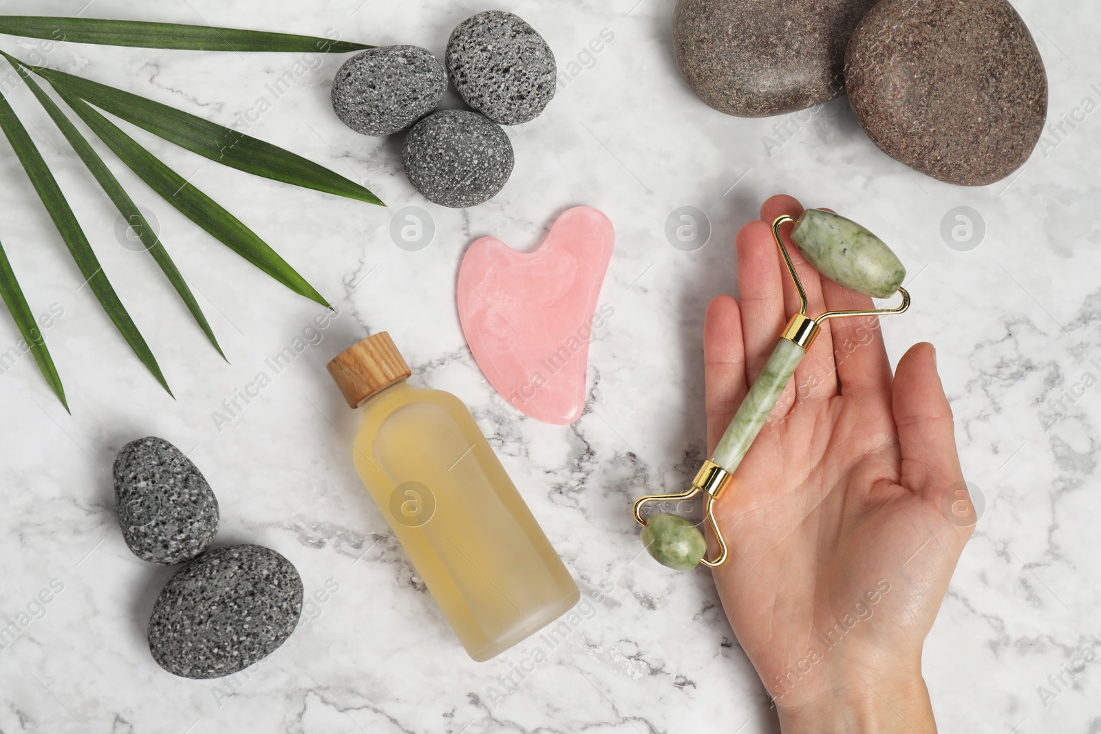 Photo of Woman with face roller, gua sha tool, cosmetic product and stones on white marble background, top view