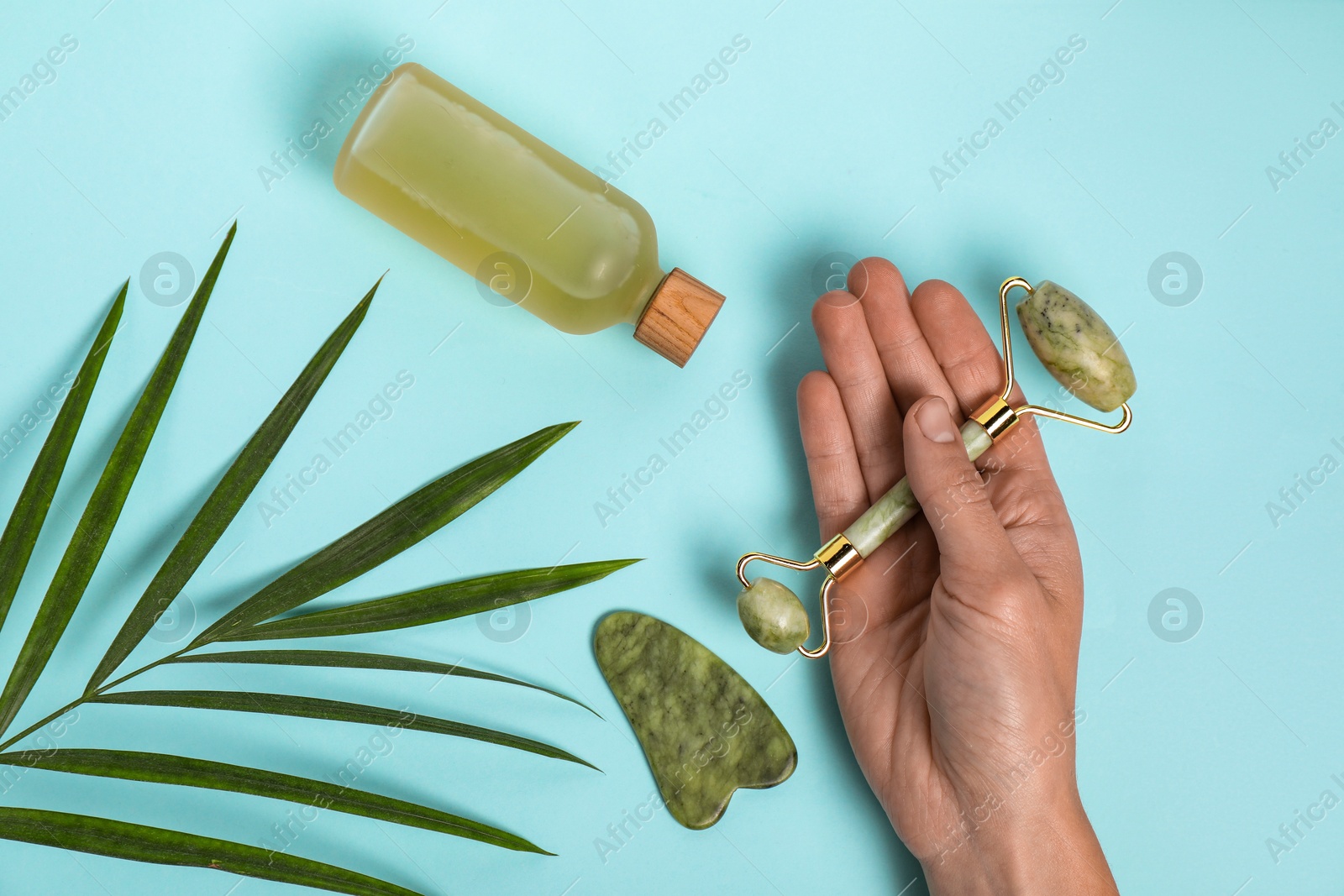 Photo of Woman with face roller, gua sha tool, cosmetic product and leaf on light blue background, top view