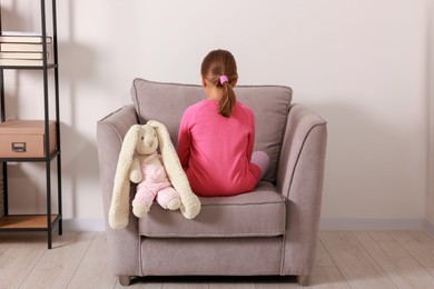 Photo of Autism concept. Lonely little girl with toy bunny in armchair at home, back view