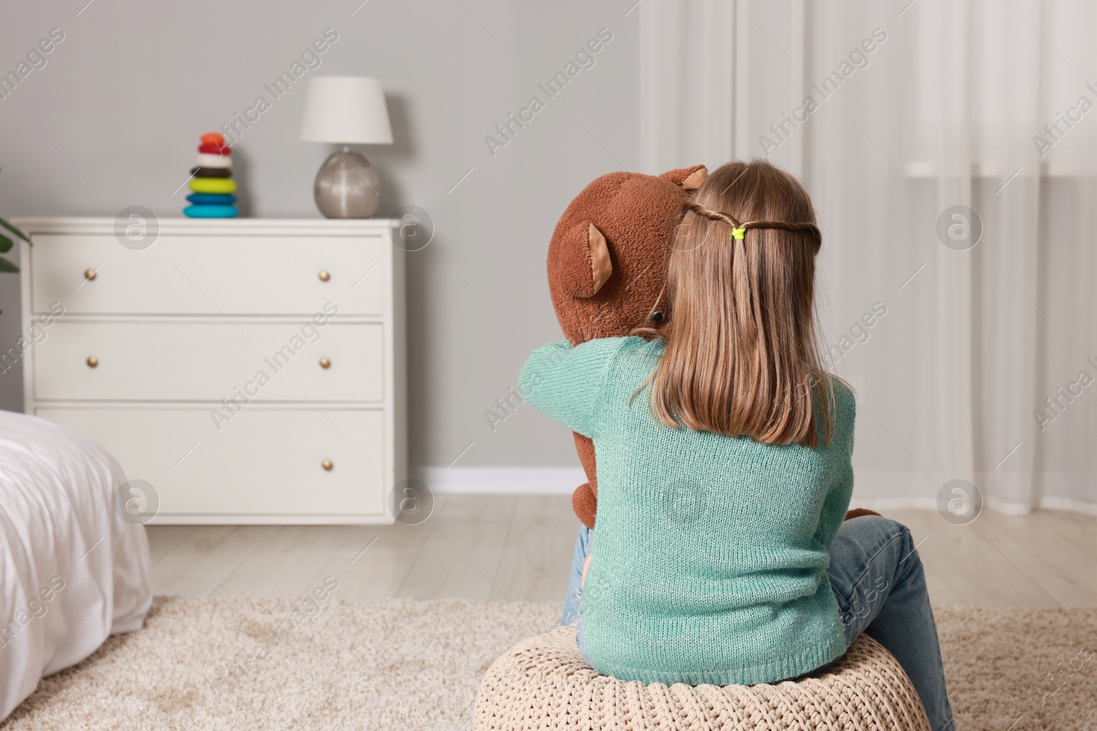 Photo of Autism concept. Lonely little girl with teddy bear on pouf at home, back view