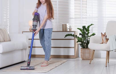 Photo of Teenage girl cleaning rug with cordless vacuum cleaner and her cute Chihuahua dog sitting in armchair at home, closeup