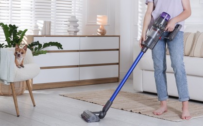 Photo of Teenage girl cleaning floor with cordless vacuum cleaner and her cute Chihuahua dog sitting in armchair at home, closeup