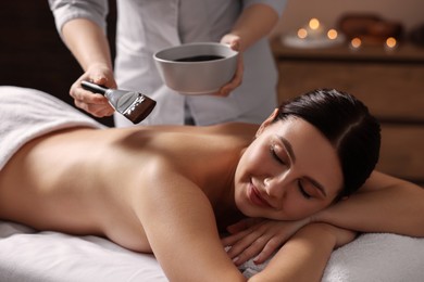 Photo of Chocolate body wrap. Spa worker applying mask onto woman's back in salon, closeup