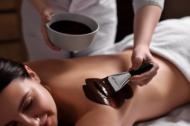 Photo of Chocolate body wrap. Spa worker applying mask onto woman's back in salon, closeup