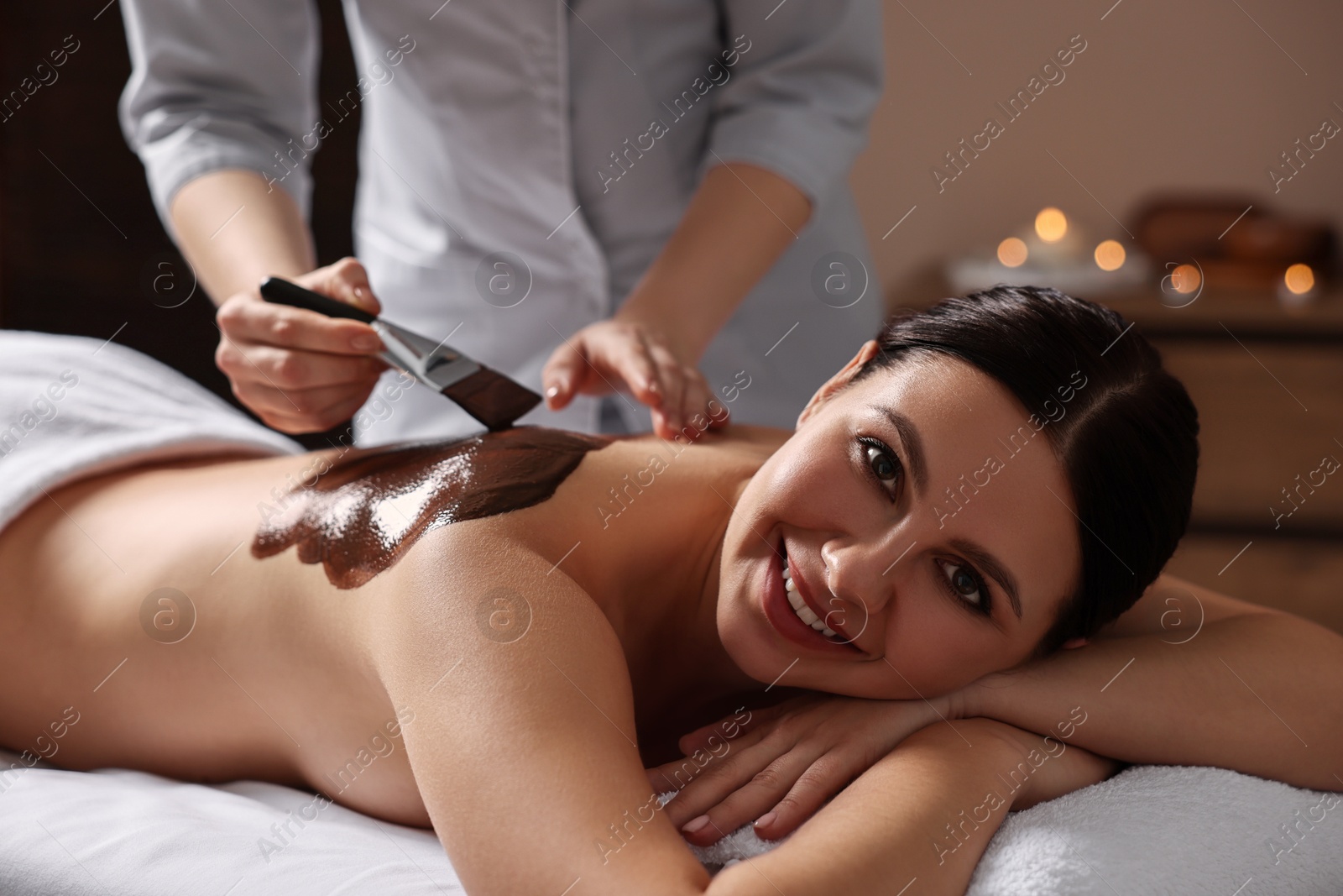 Photo of Chocolate body wrap. Spa worker applying mask onto woman's back in salon, closeup