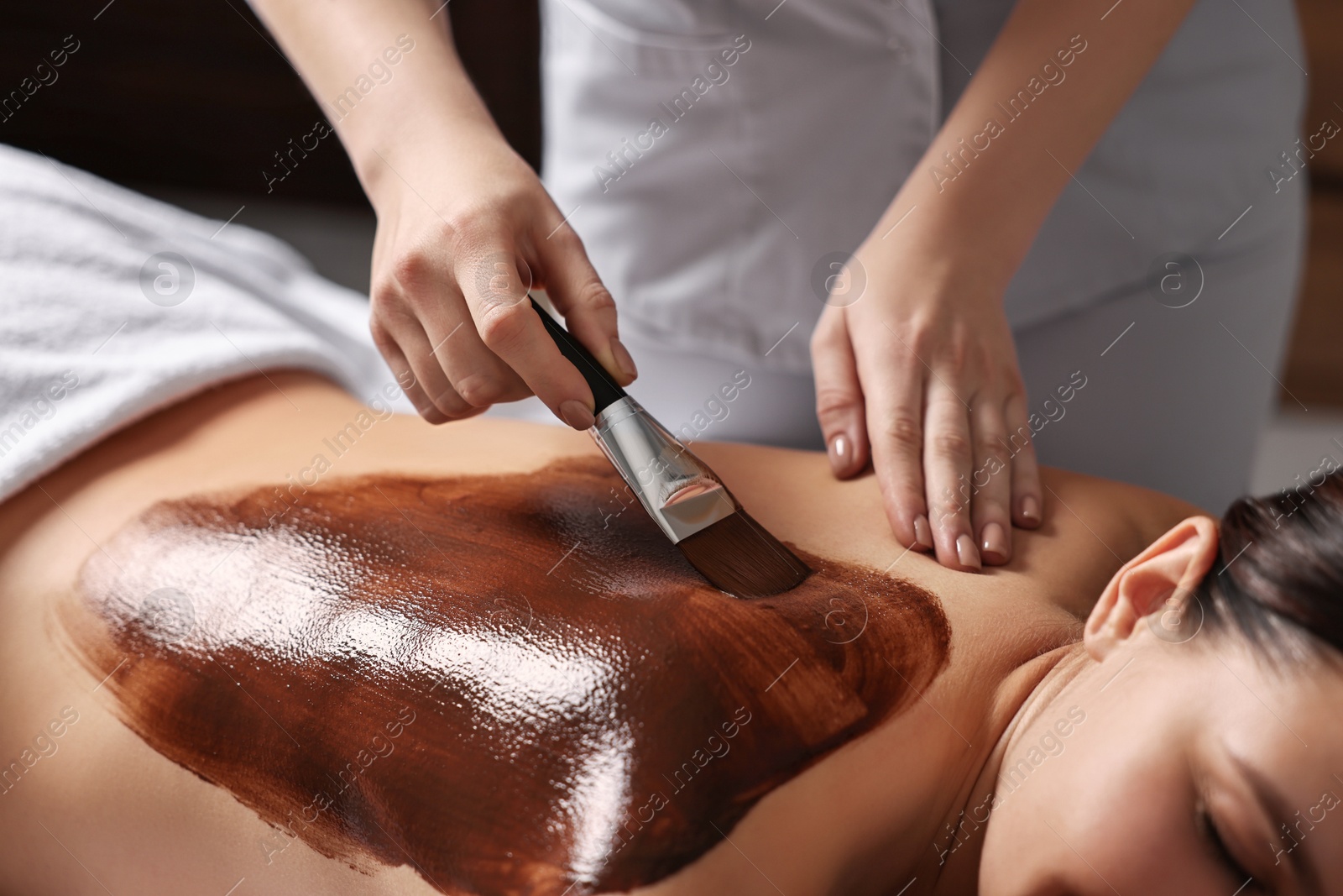 Photo of Chocolate body wrap. Spa worker applying mask onto woman's back in salon, closeup