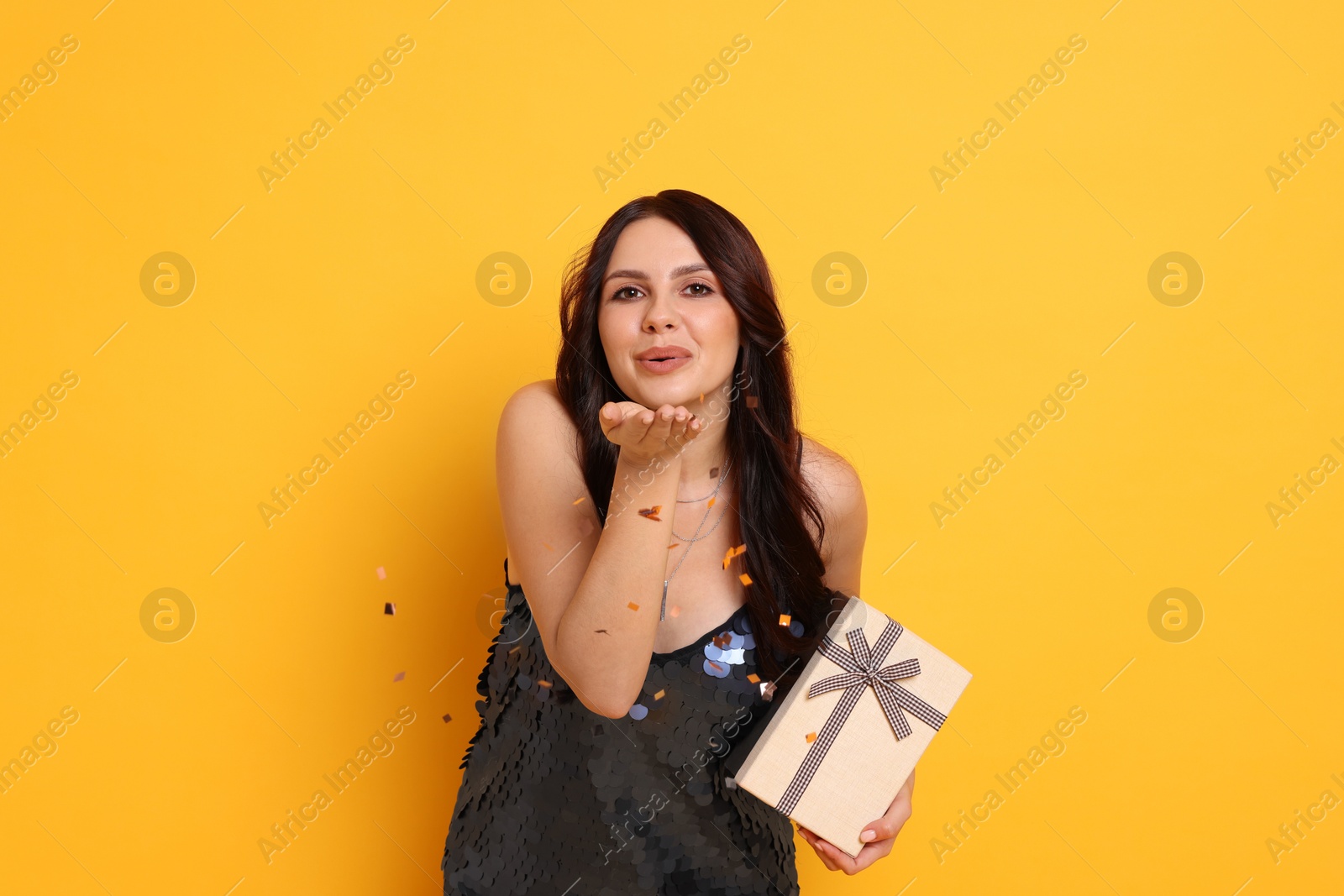 Photo of Beautiful woman with gift box blowing confetti off hands on orange background. Surprise party