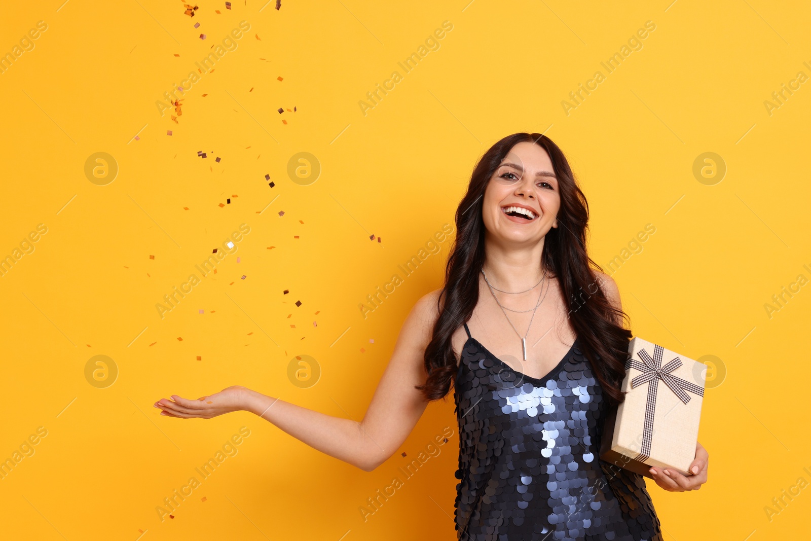 Photo of Happy woman with gift box and flying confetti on orange background. Surprise party