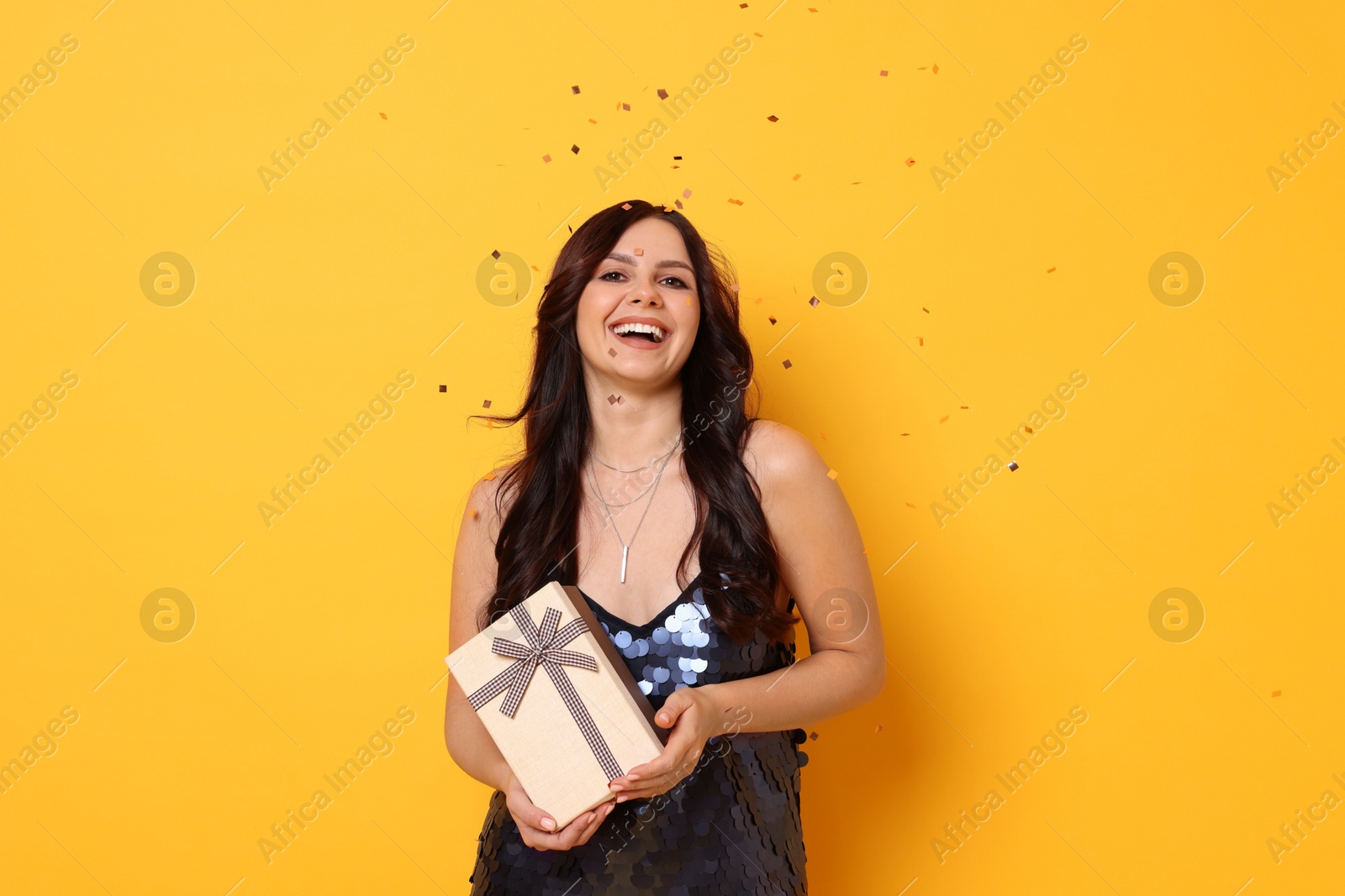 Photo of Happy woman with gift box under falling confetti on orange background. Surprise party