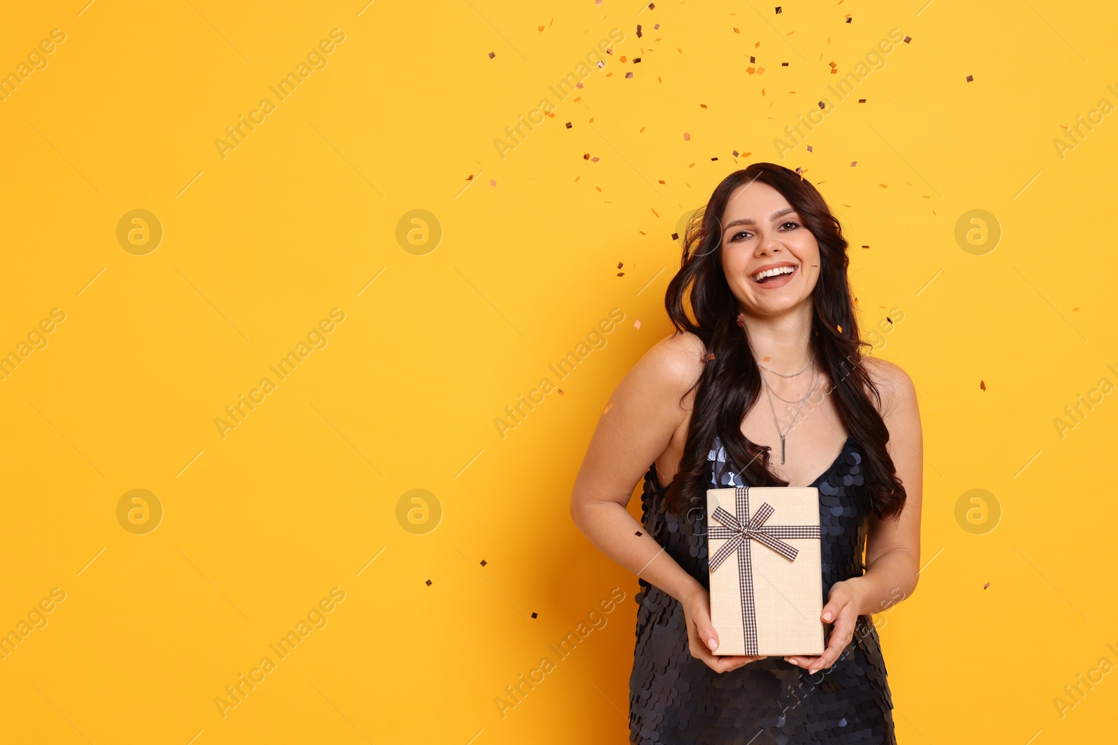 Photo of Happy woman with gift box under falling confetti on orange background, space for text. Surprise party