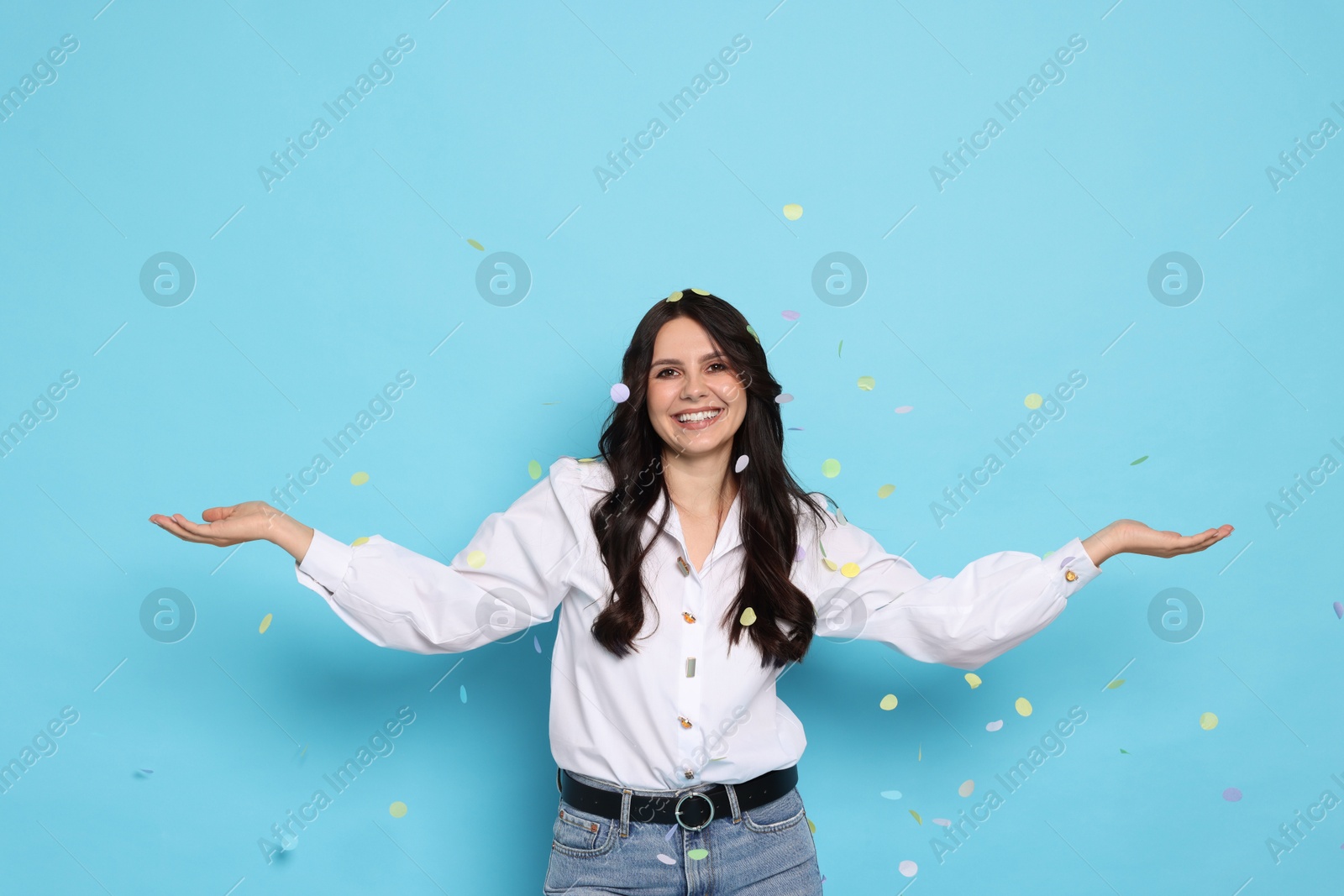 Photo of Happy woman under falling confetti on light blue background. Surprise party