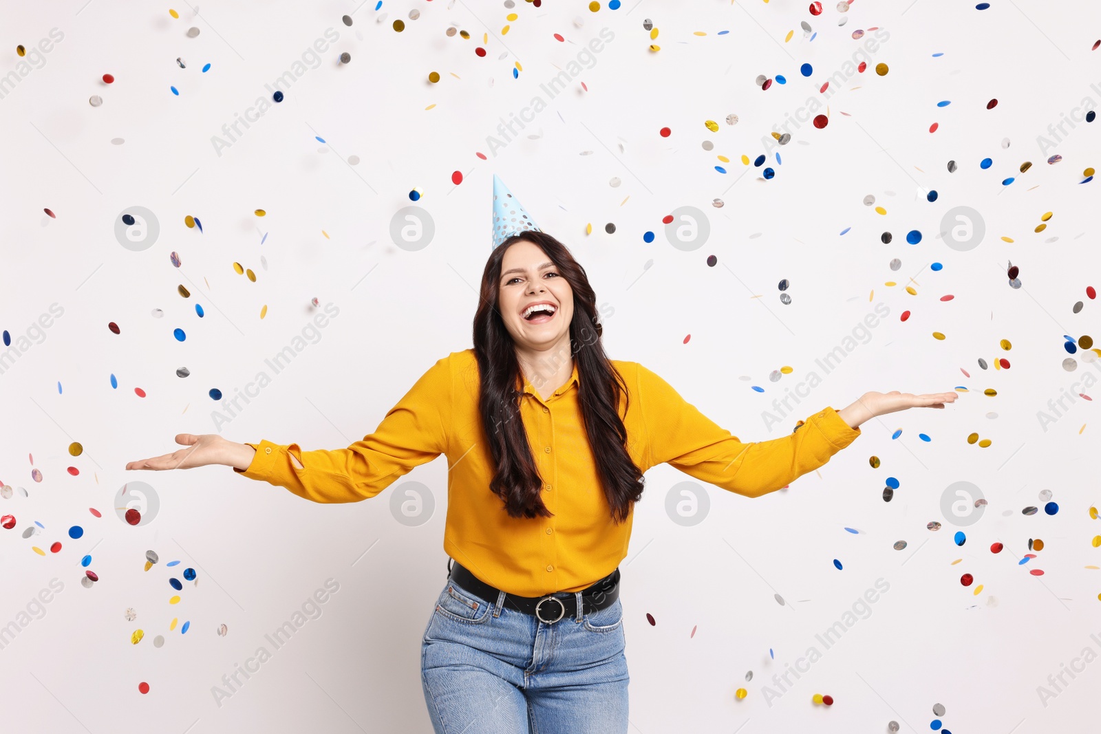 Photo of Happy woman in conical paper hat under falling confetti on white background. Surprise party