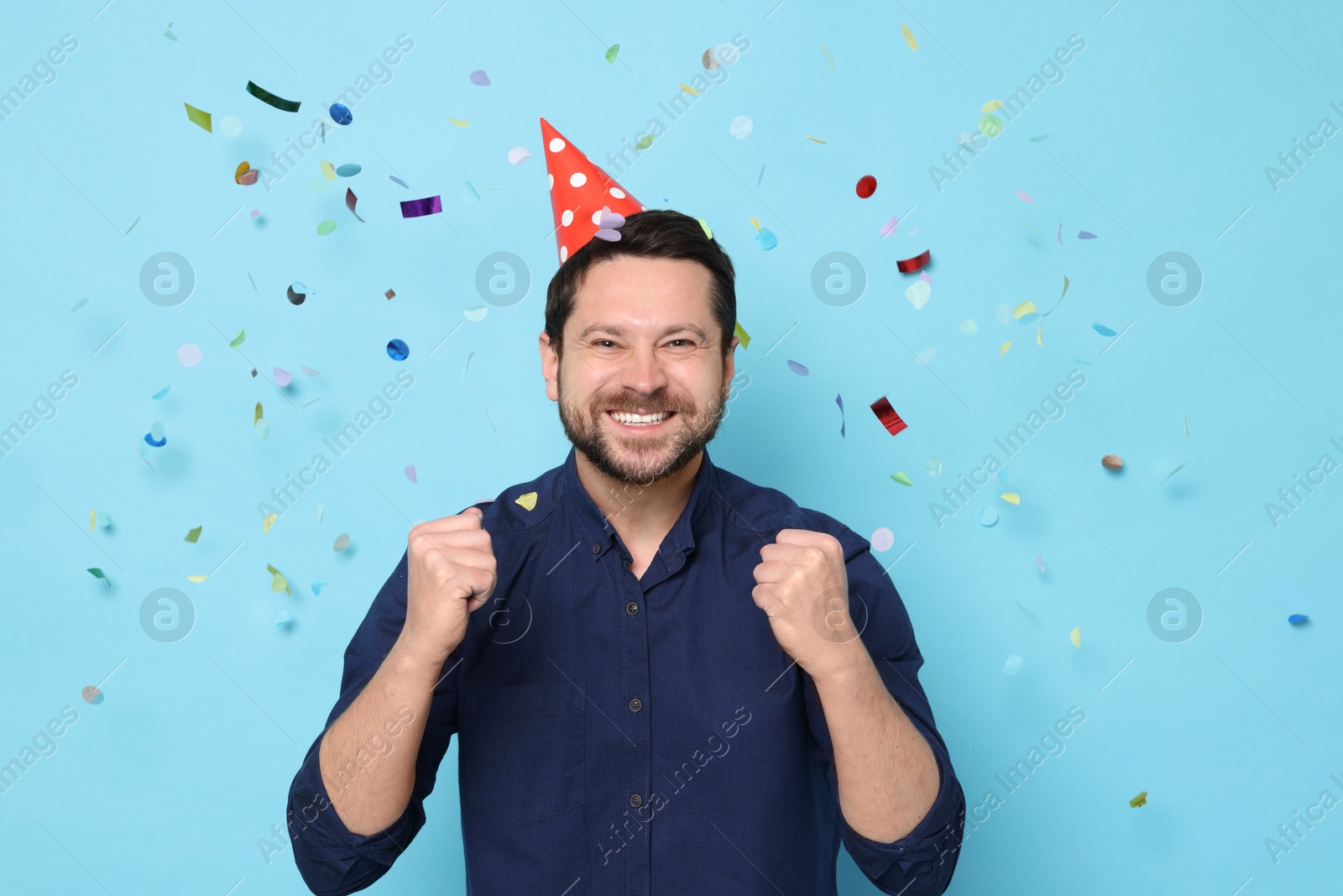 Photo of Happy man in conical paper hat under falling confetti on light blue background. Surprise party