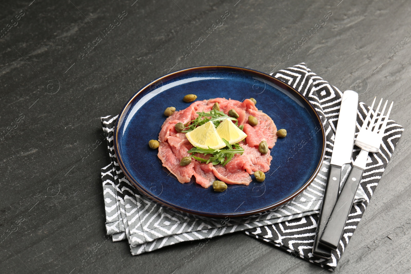 Photo of Fresh beef carpaccio with arugula, lemon and capers on dark textured table