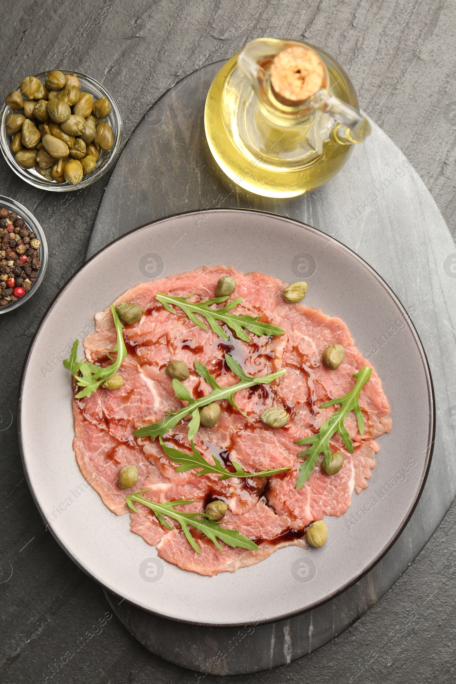 Photo of Fresh beef carpaccio with arugula, capers and sauce on dark textured table, flat lay