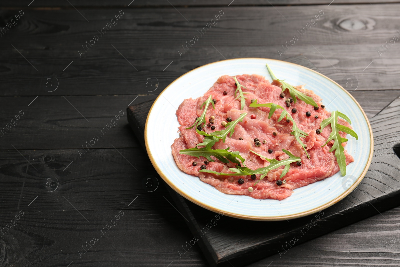 Photo of Fresh beef carpaccio with arugula and peppercorns on black wooden table, closeup. Space for text