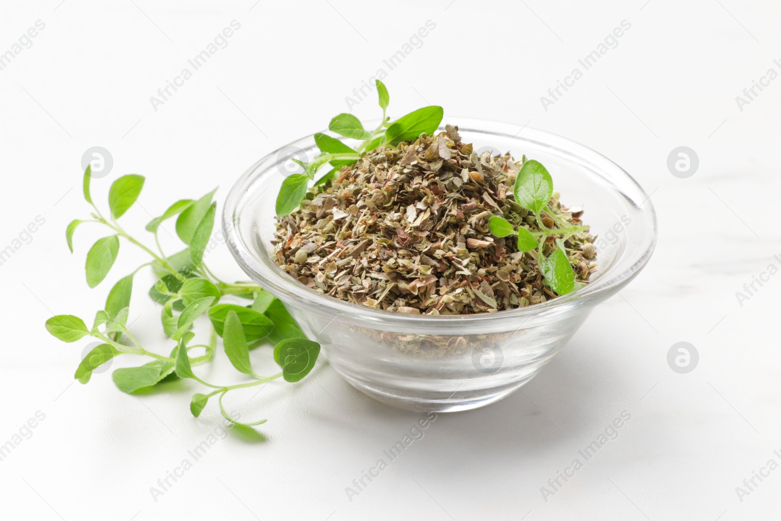 Photo of Dried oregano in glass bowl and green leaves on white table, closeup