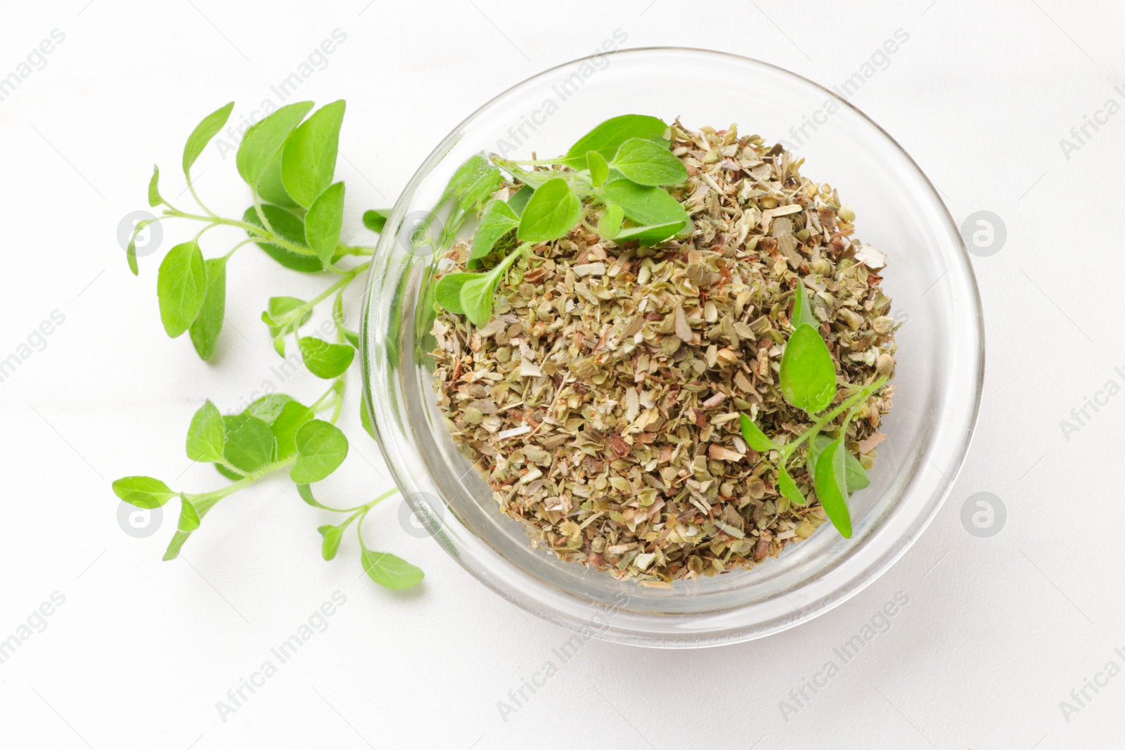 Photo of Dried oregano in glass bowl and green leaves on white table, top view