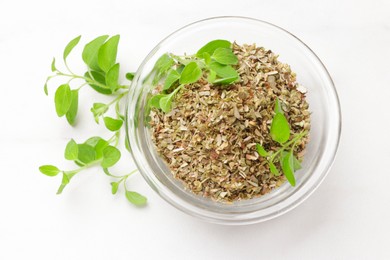 Dried oregano in glass bowl and green leaves on white table, top view