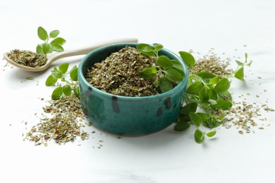 Dried oregano in bowl, spoon and green leaves on white table