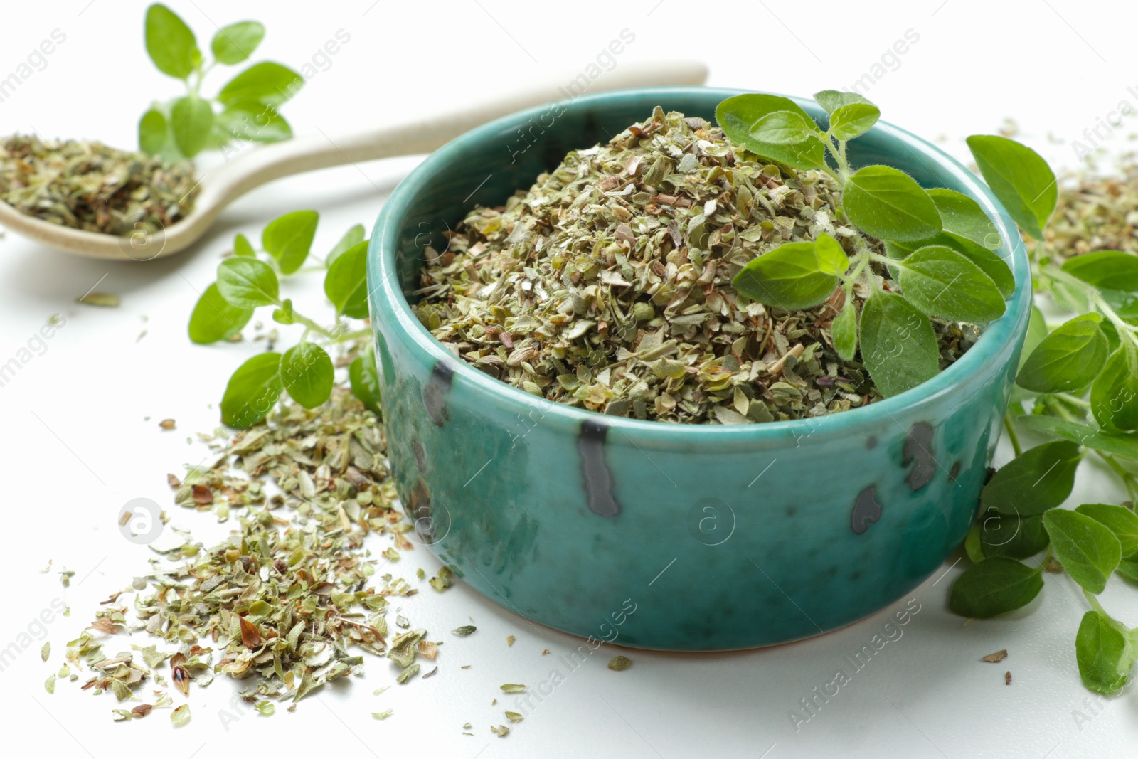 Photo of Dried oregano in bowl, spoon and green leaves on white table, closeup