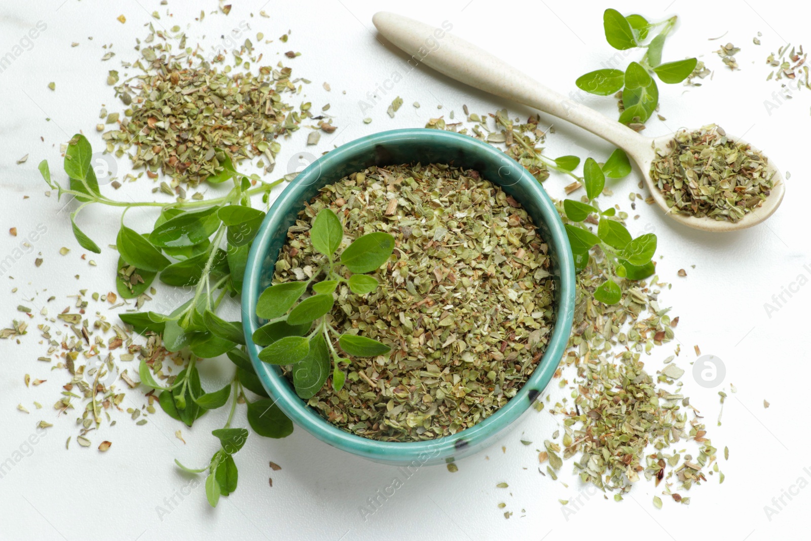 Photo of Dried oregano in bowl, spoon and green leaves on white table, top view