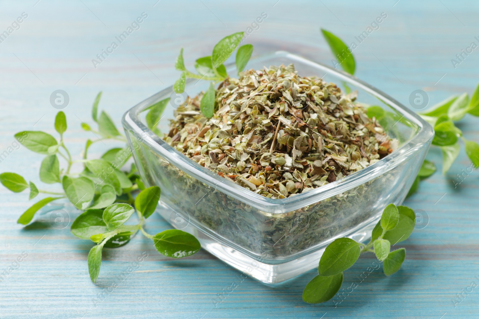 Photo of Dried oregano in glass bowl and green leaves on light blue wooden table, closeup