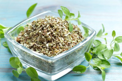 Photo of Dried oregano in glass bowl and green leaves on light blue wooden table, closeup