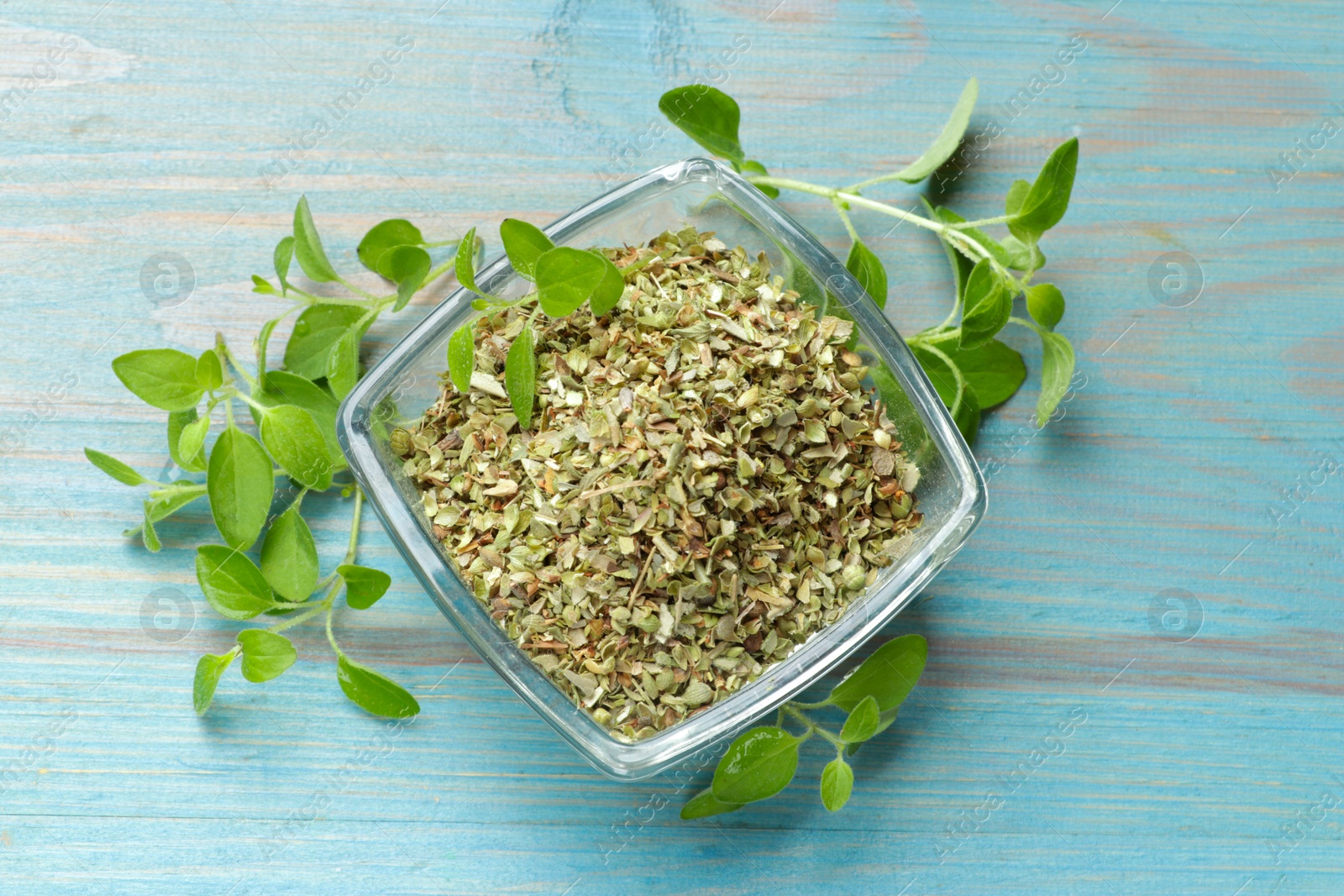 Photo of Dried oregano in glass bowl and green leaves on light blue wooden table, top view