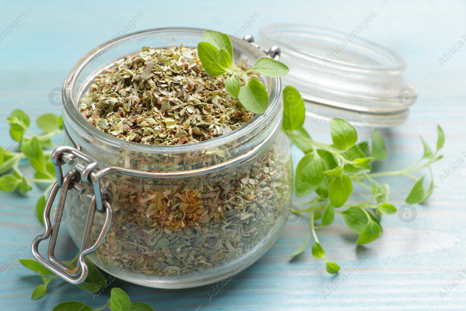 Photo of Dried oregano in glass jar and green leaves on light blue wooden table, closeup