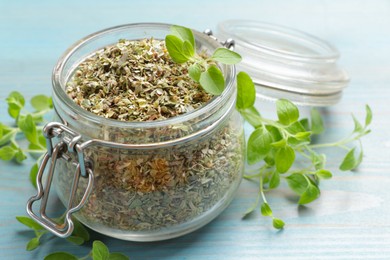 Photo of Dried oregano in glass jar and green leaves on light blue wooden table, closeup