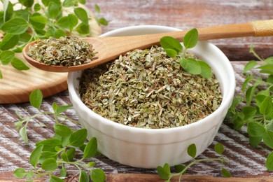 Photo of Dried oregano in bowl, spoon and green leaves on wooden table, closeup