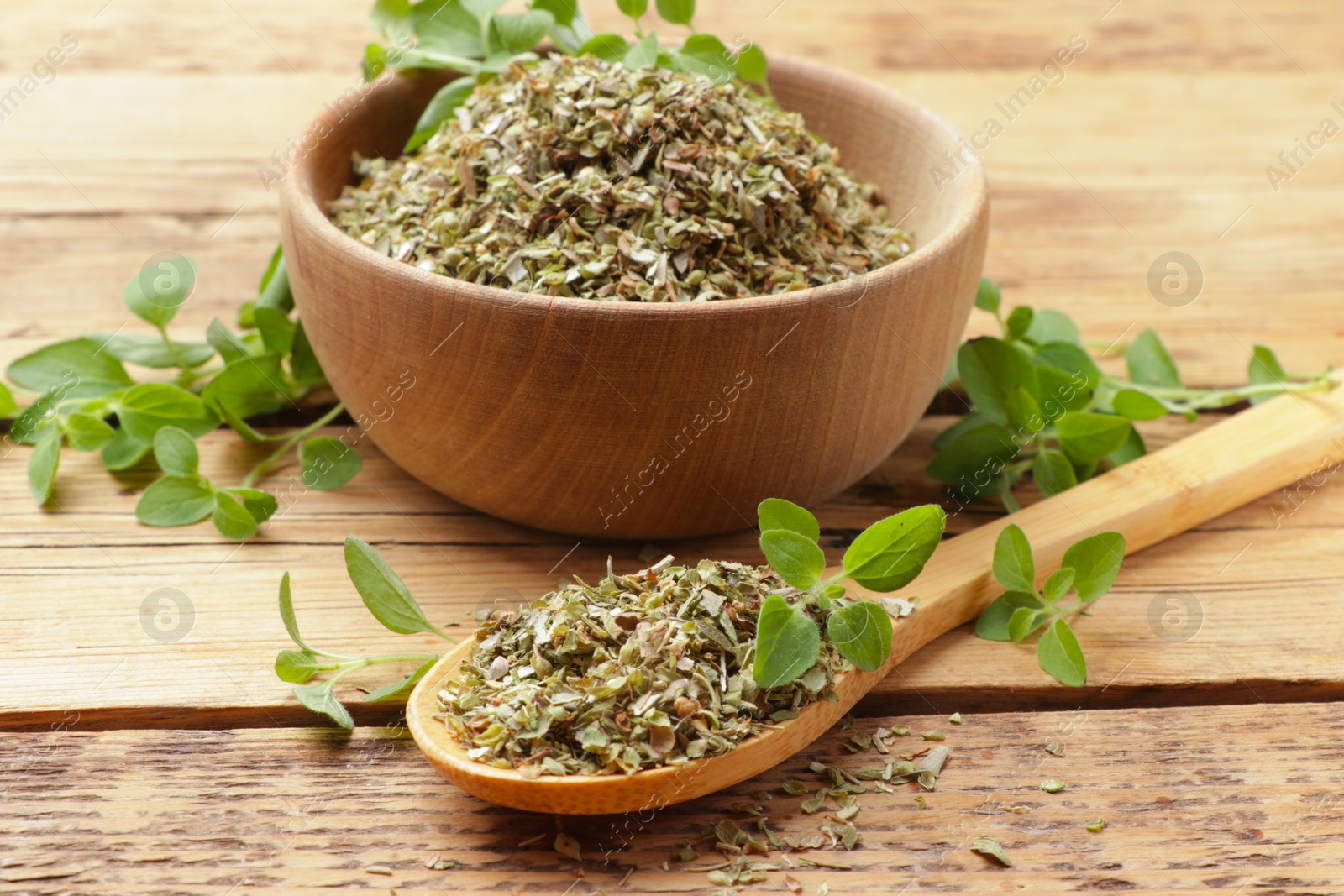 Photo of Dried oregano in bowl, spoon and green leaves on wooden table, closeup