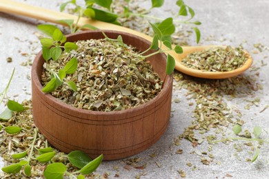 Dried oregano in bowl, spoon and green leaves on light grey table, closeup