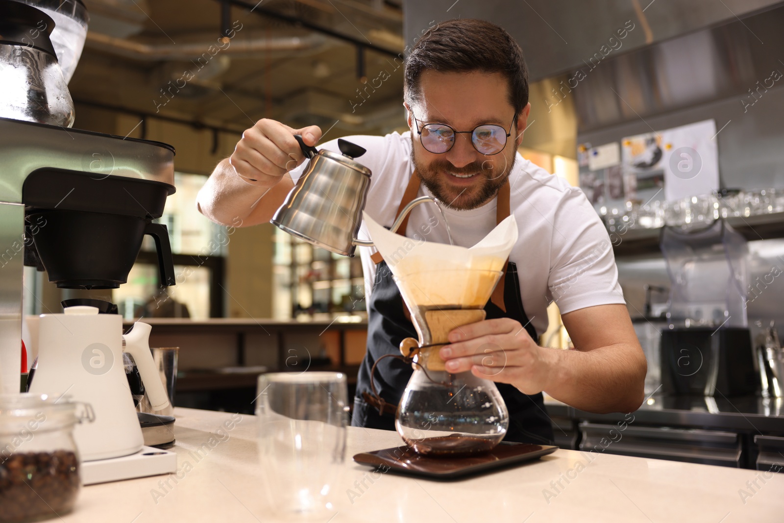 Photo of Barista brewing coffee in glass coffeemaker with paper filter at table in cafe