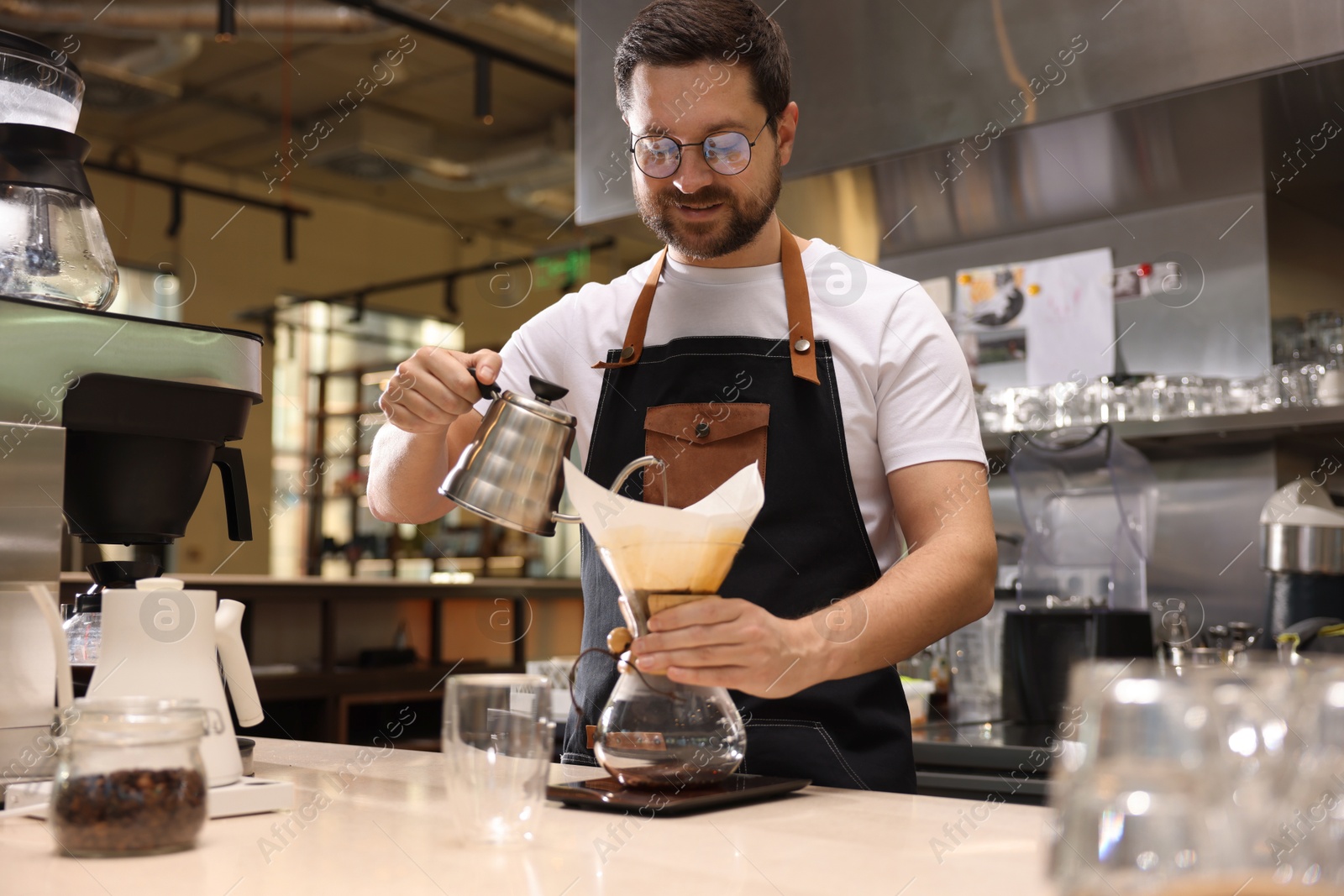 Photo of Barista brewing coffee in glass coffeemaker with paper filter at table in cafe