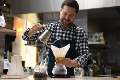 Photo of Barista brewing coffee in glass coffeemaker with paper filter at table in cafe