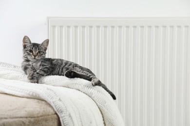 Photo of Cute little kitten on pouf near radiator at home, space for text