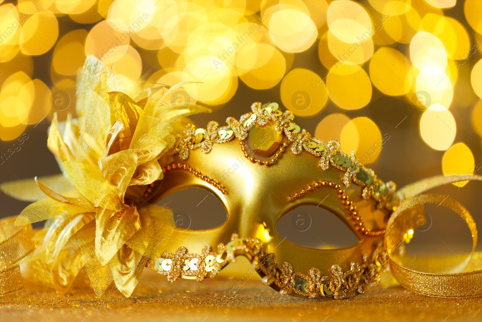 Photo of Beautiful carnival mask on golden shiny surface against blurred lights, closeup