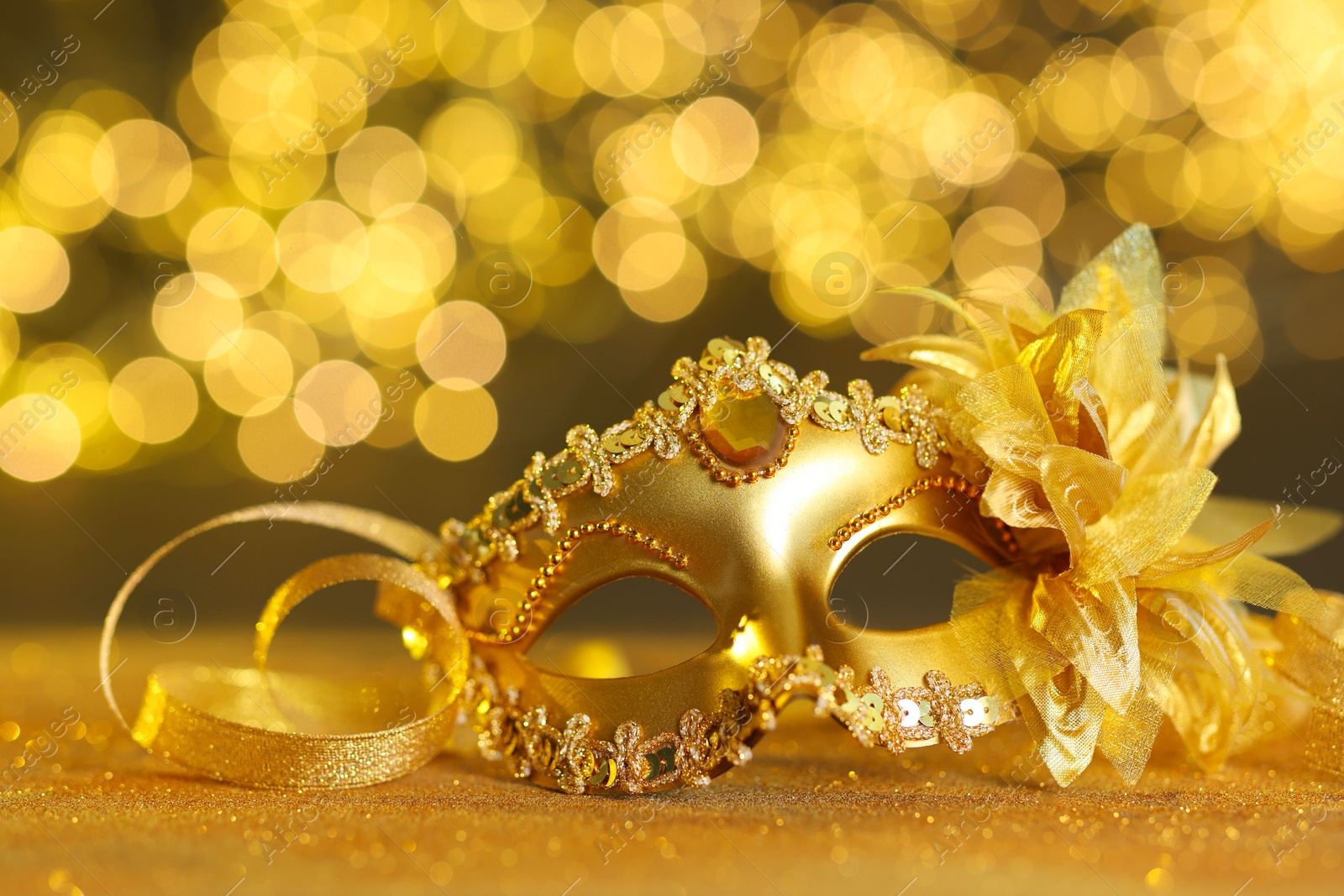 Photo of Beautiful carnival mask on golden shiny surface against blurred lights, closeup