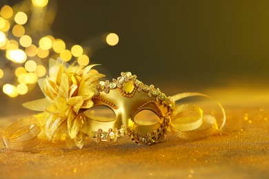 Photo of Beautiful carnival mask on golden shiny surface against blurred lights, space for text