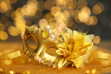 Photo of Beautiful carnival mask on golden shiny surface against blurred lights, closeup