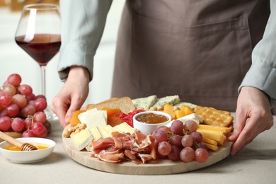 Photo of Woman holding board with different types of cut cheese and other snacks at light textured table, closeup