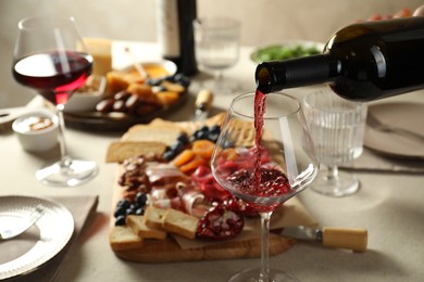 Photo of Pouring red wine into glass at light grey table with different snacks, closeup