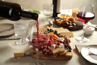 Photo of Pouring red wine into glass at light grey table with different snacks, closeup