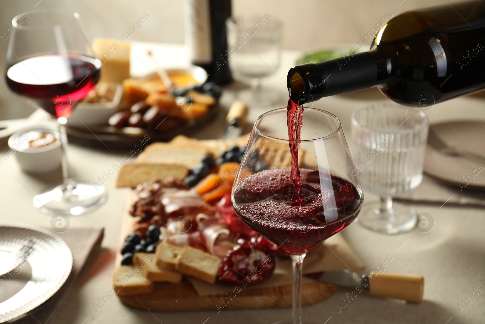 Photo of Pouring red wine into glass at light grey table with different snacks, closeup