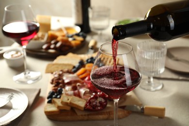 Photo of Pouring red wine into glass at light grey table with different snacks, closeup