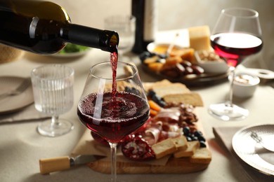 Photo of Pouring red wine into glass at light grey table with different snacks, closeup