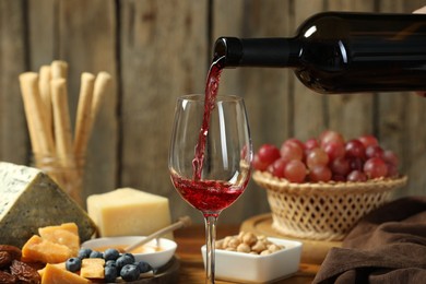 Photo of Pouring red wine into glass at table with different snacks, closeup